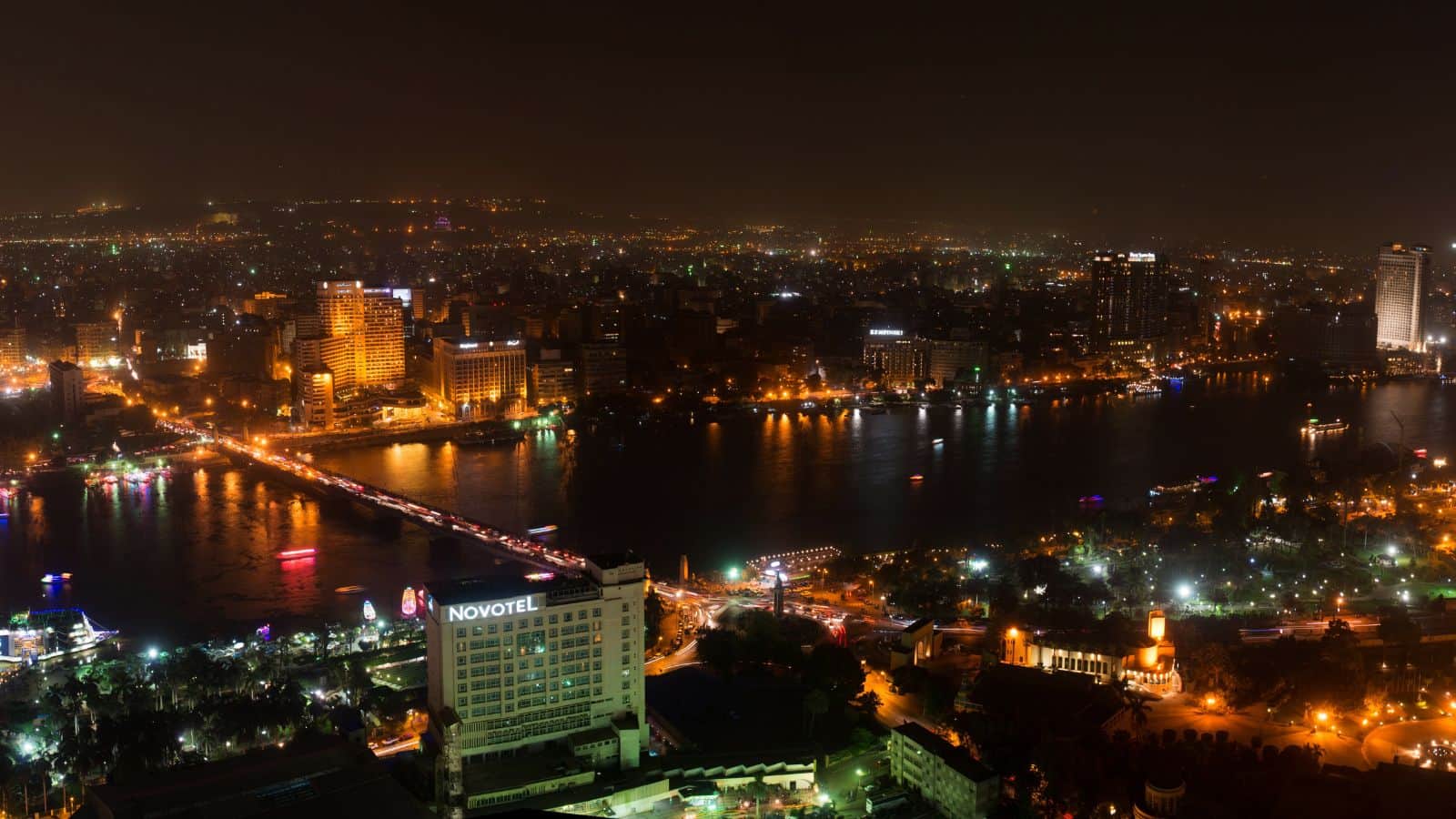 Aerial view of a city at night with a river running through the center. The cityscape is illuminated with numerous lights from buildings and streets. A bridge crosses the river, and a hotel named "Novotel" is visible in the foreground.