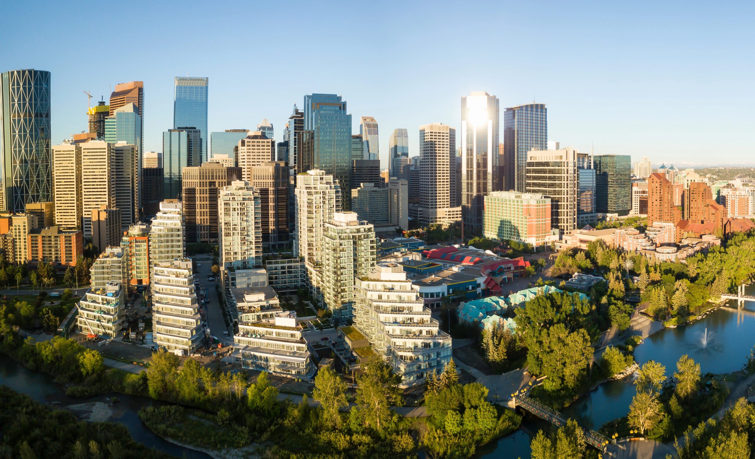 Aerial view of a city skyline with high-rises, residential areas, green spaces, and a river under a clear blue sky.