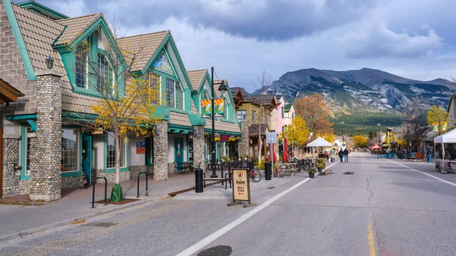 A colorful shop-lined street with autumn trees, set against mountains and a partly cloudy sky.