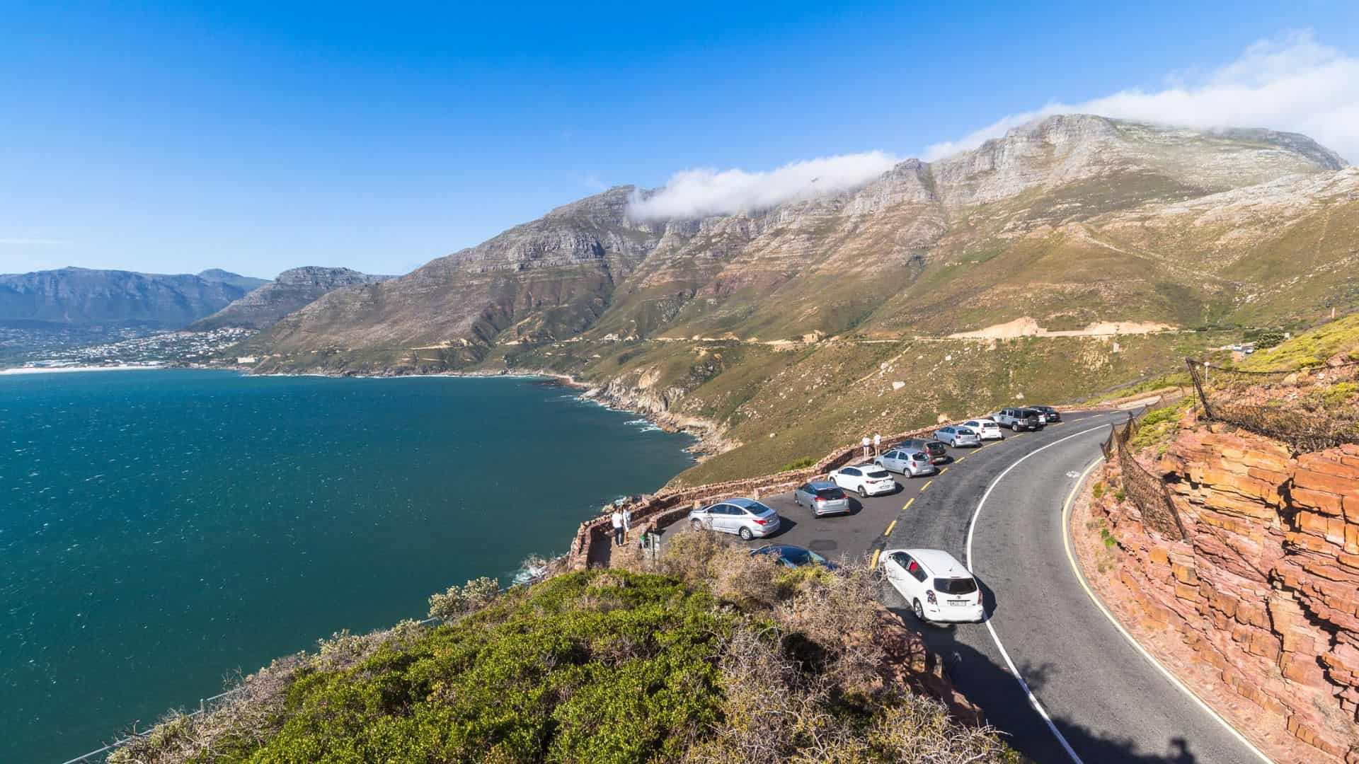 Cars line a coastal road overlooking blue ocean and distant mountains beneath a clear sky.
