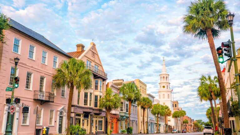 Colorful historic buildings and palm trees line a street beneath a blue sky, with a white church steeple in the background.
