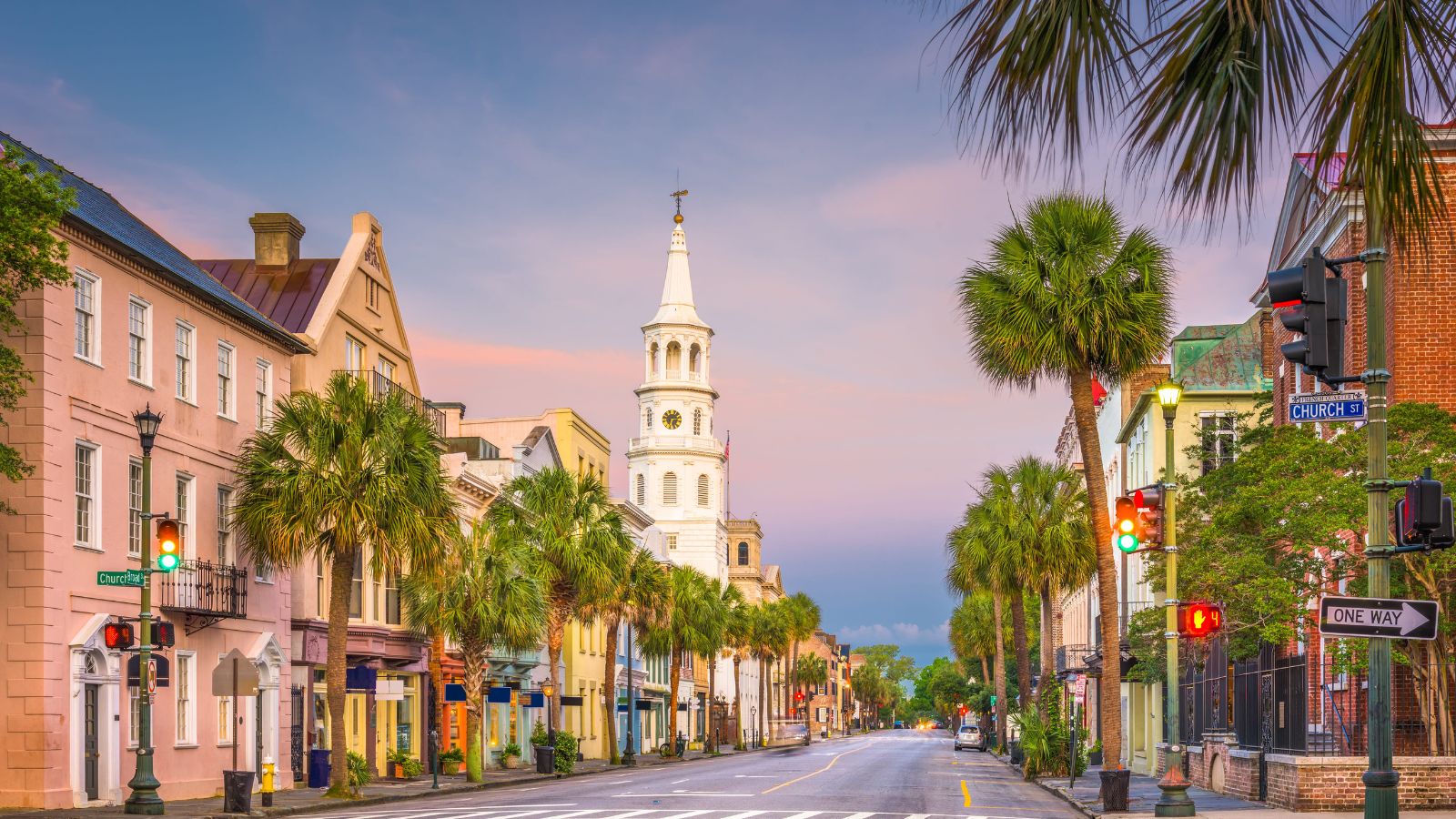 A Charleston street at sunset with colorful historic buildings, palm trees, and a white church steeple in the background.