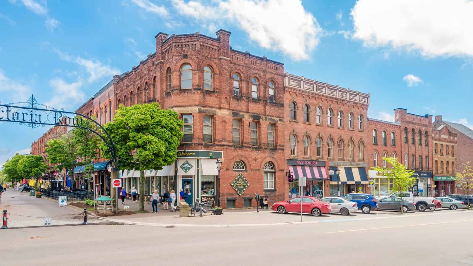 Brick buildings, shops, parked cars, and pedestrians line a historic downtown street beneath a blue sky with clouds.