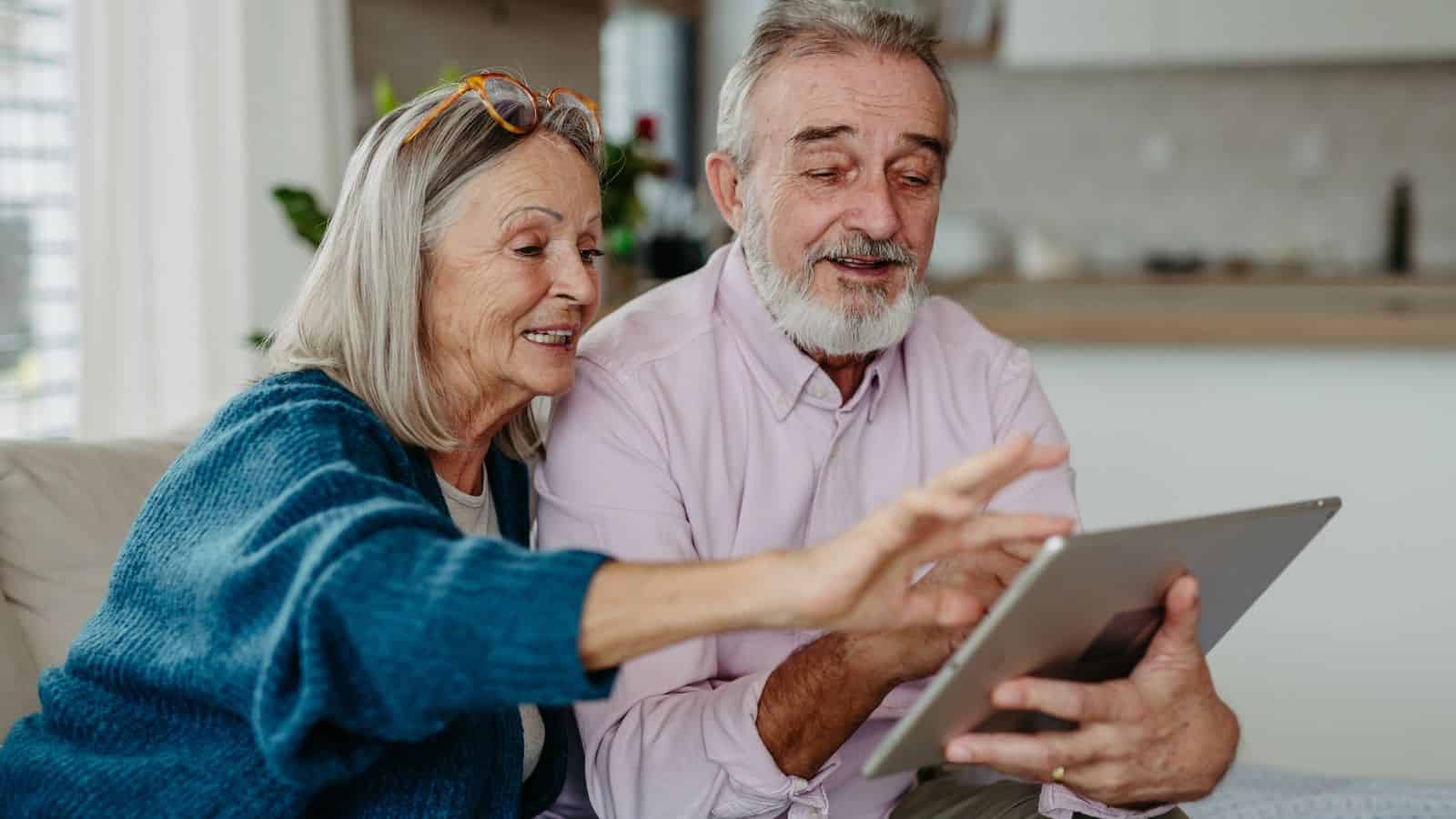 An older man and woman smile while looking at a tablet together on a couch in a bright living room.