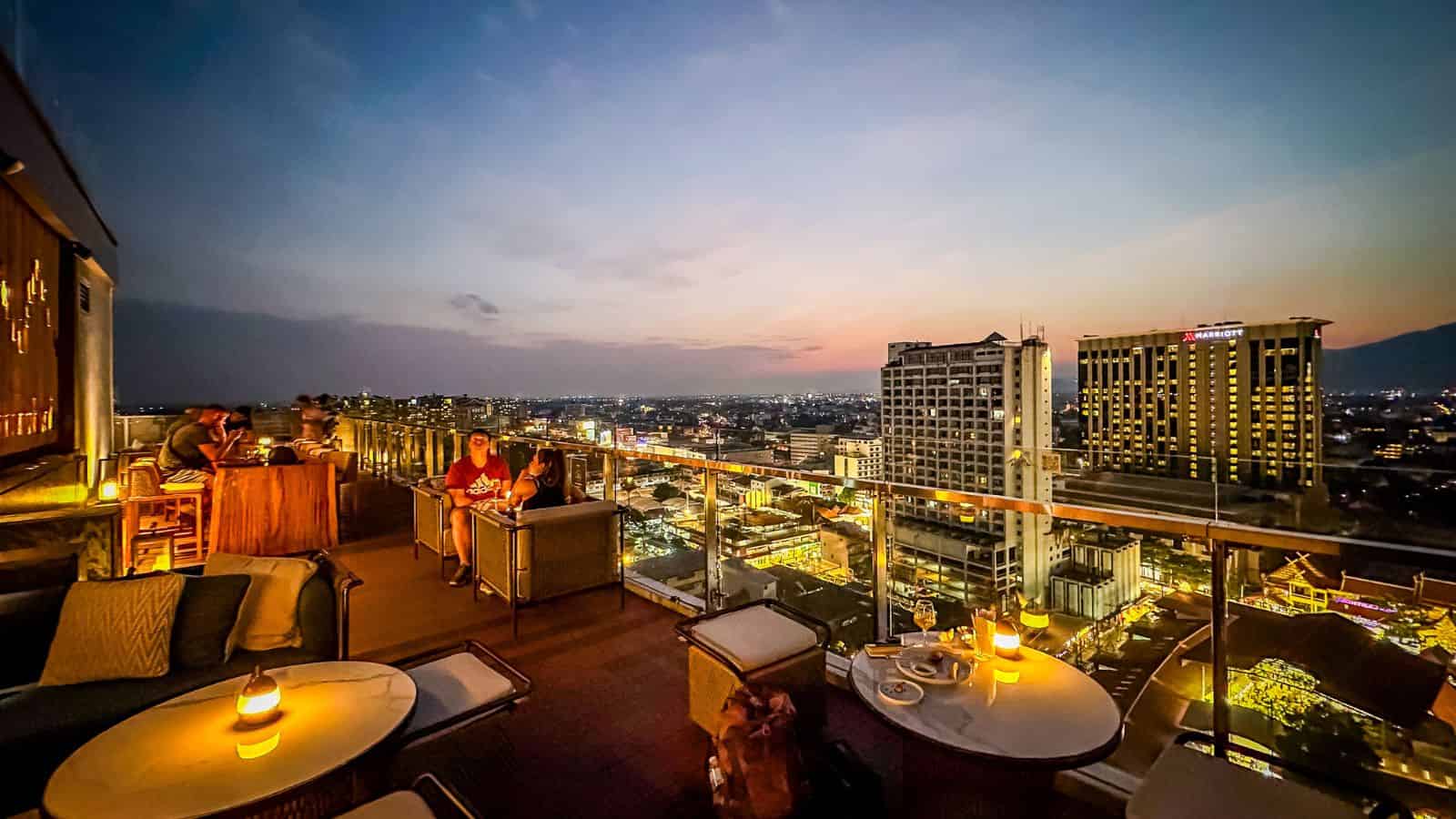 People sit at tables on a rooftop bar overlooking a sunset-lit cityscape with distant buildings and lights.
