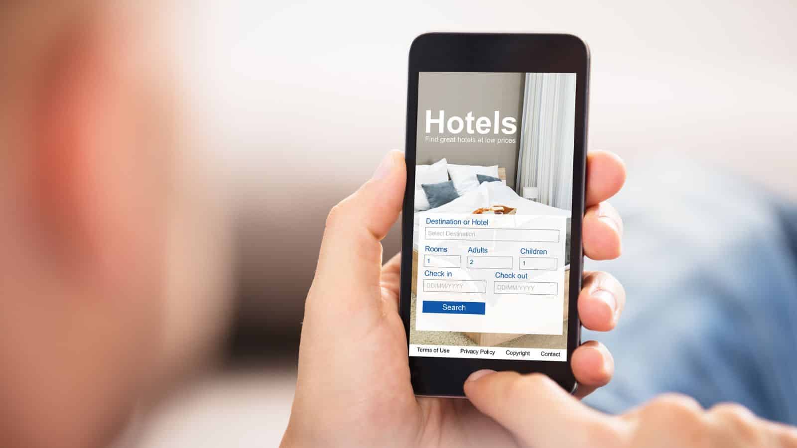 A person holds a smartphone showing a hotel booking app with entry fields for destination, rooms, guests, and stay dates.