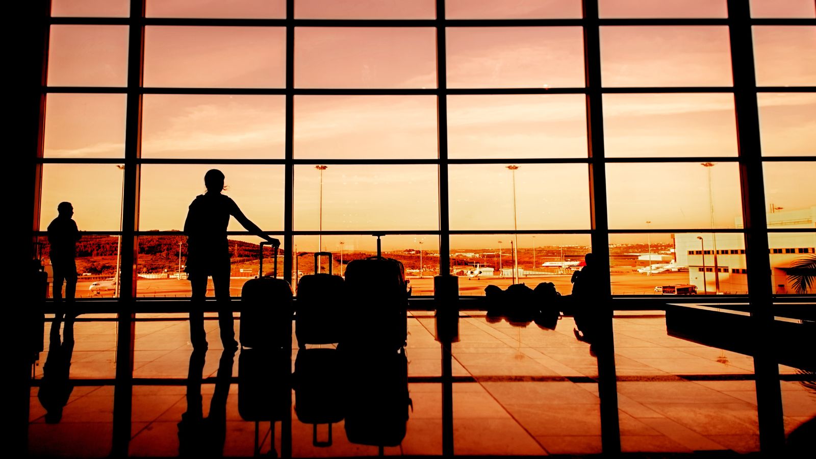 People with luggage stand silhouetted by airport windows at sunset, with planes seen on the tarmac outside.