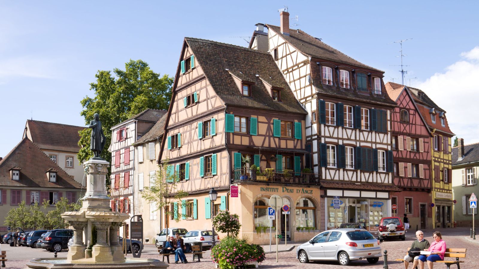 A sunny European street with colorful timber-framed buildings, parked cars, a stone statue, and people on benches.