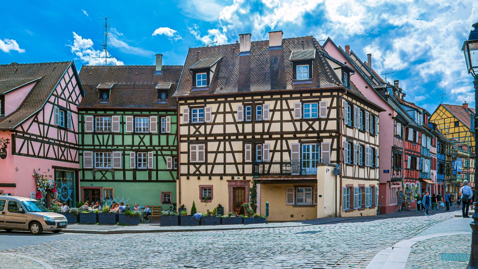 Colorful half-timbered houses border a cobblestone street with a parked car and flower planters under a partly cloudy sky.
