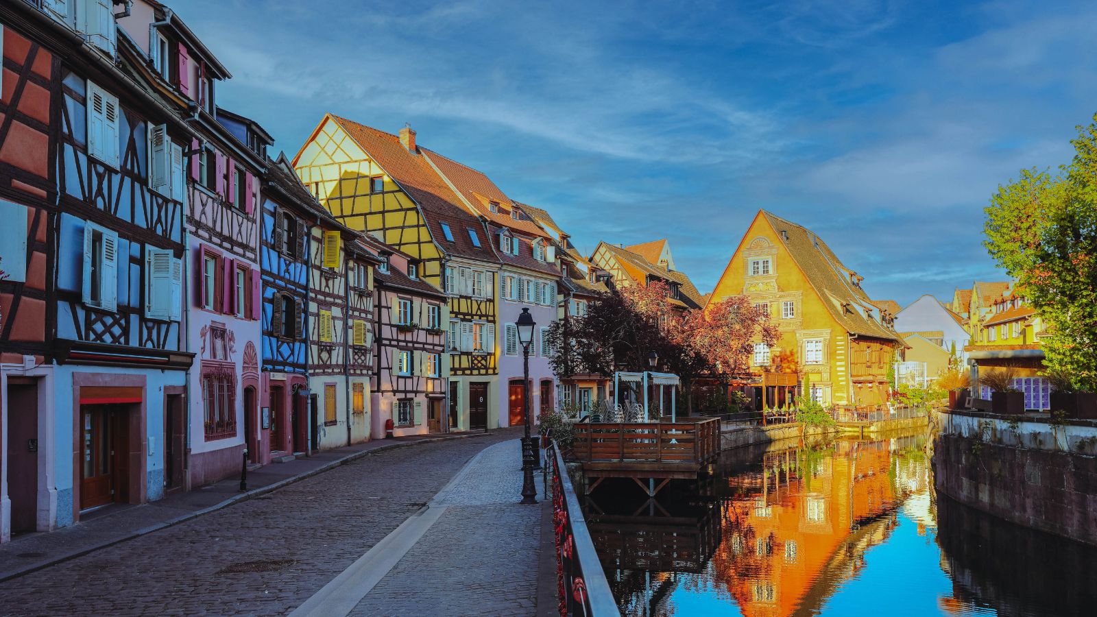 Colorful, half-timbered houses and a calm canal with building reflections line a cobblestone street in a quaint European town.