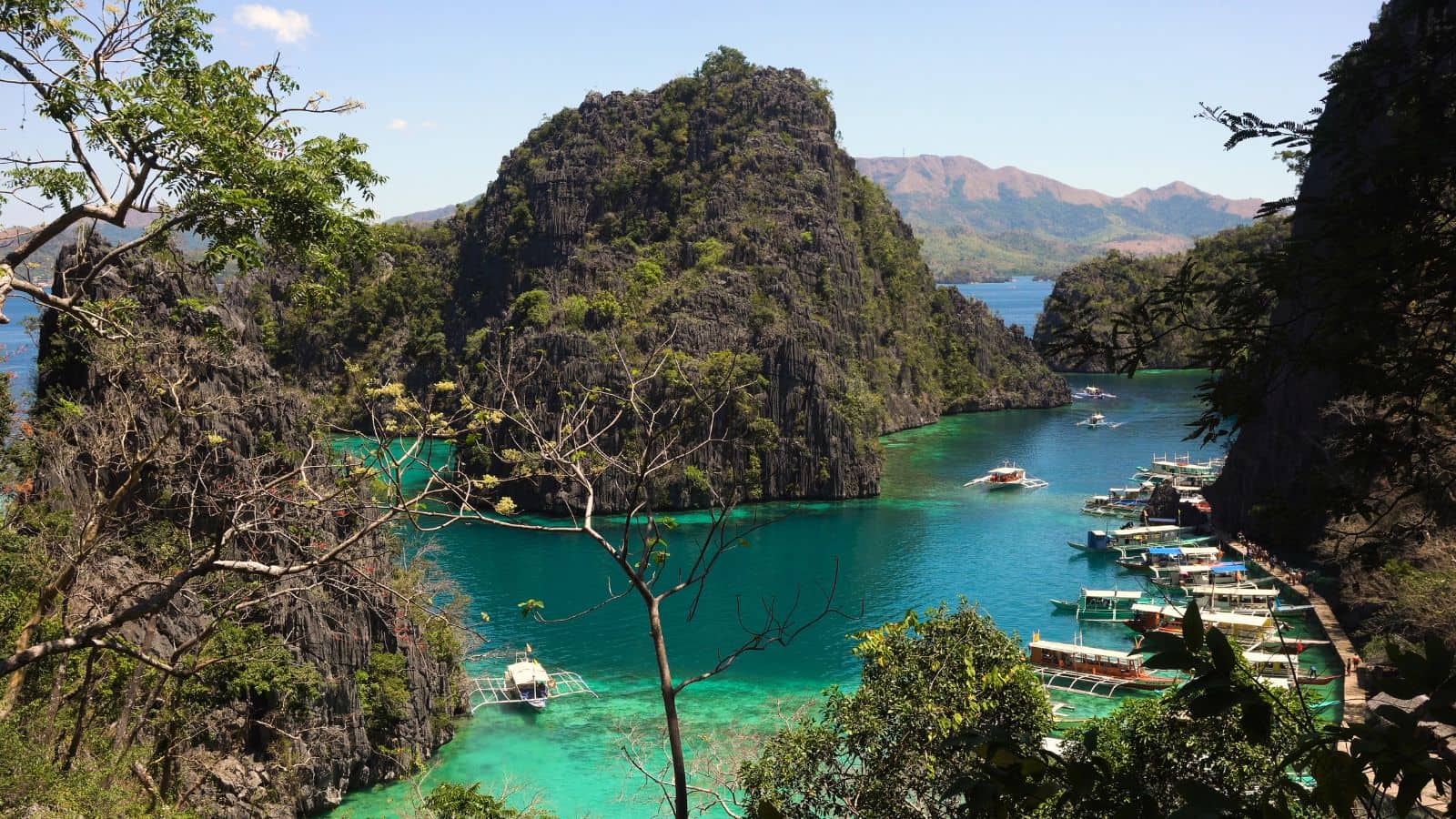 Turquoise lagoon bordered by rocky hills and greenery, with boats docked along the shore beneath a sunny sky.