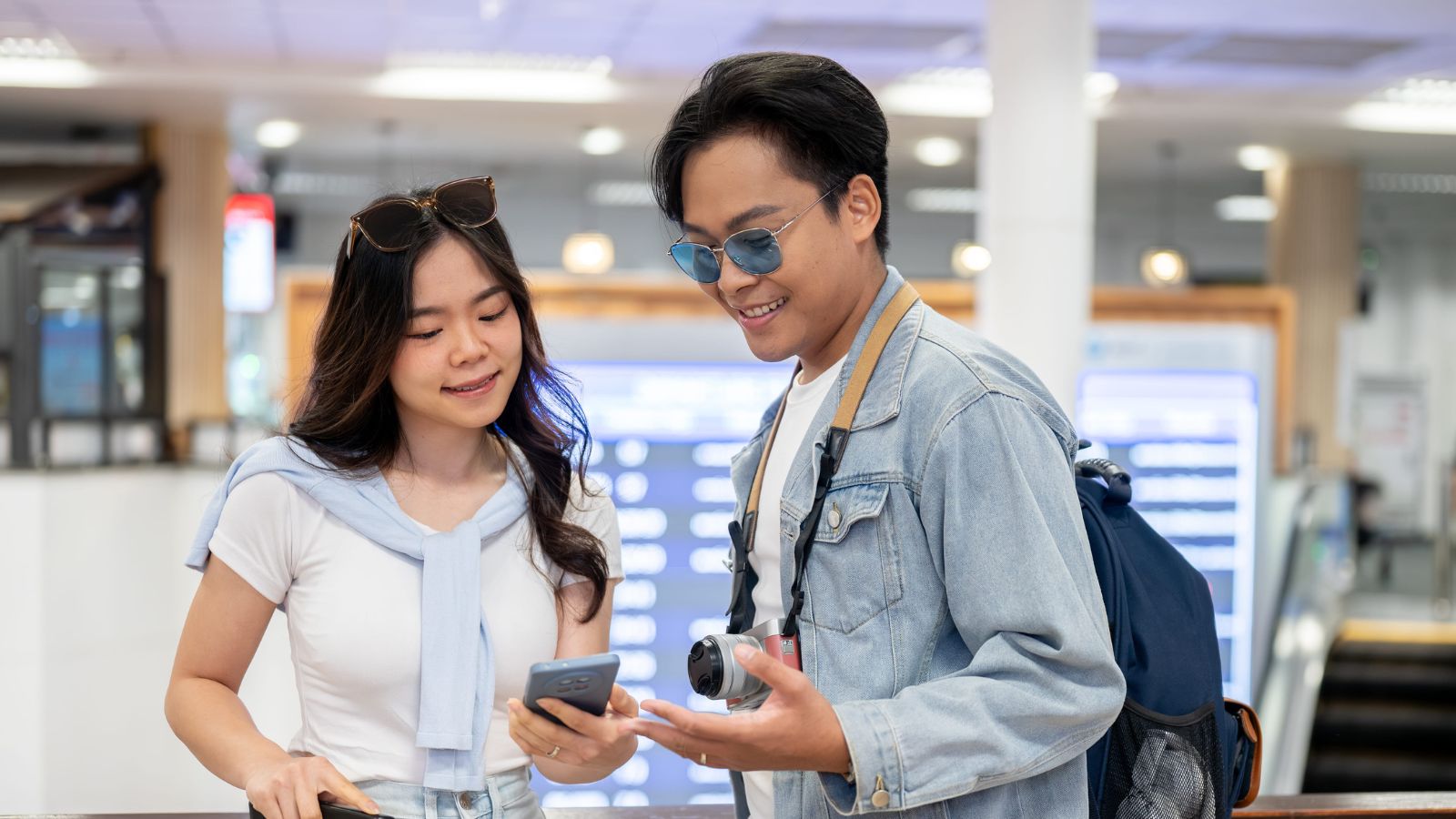 Two young adults at an airport check a smartphone, with one holding a camera and flight info screens in the background.