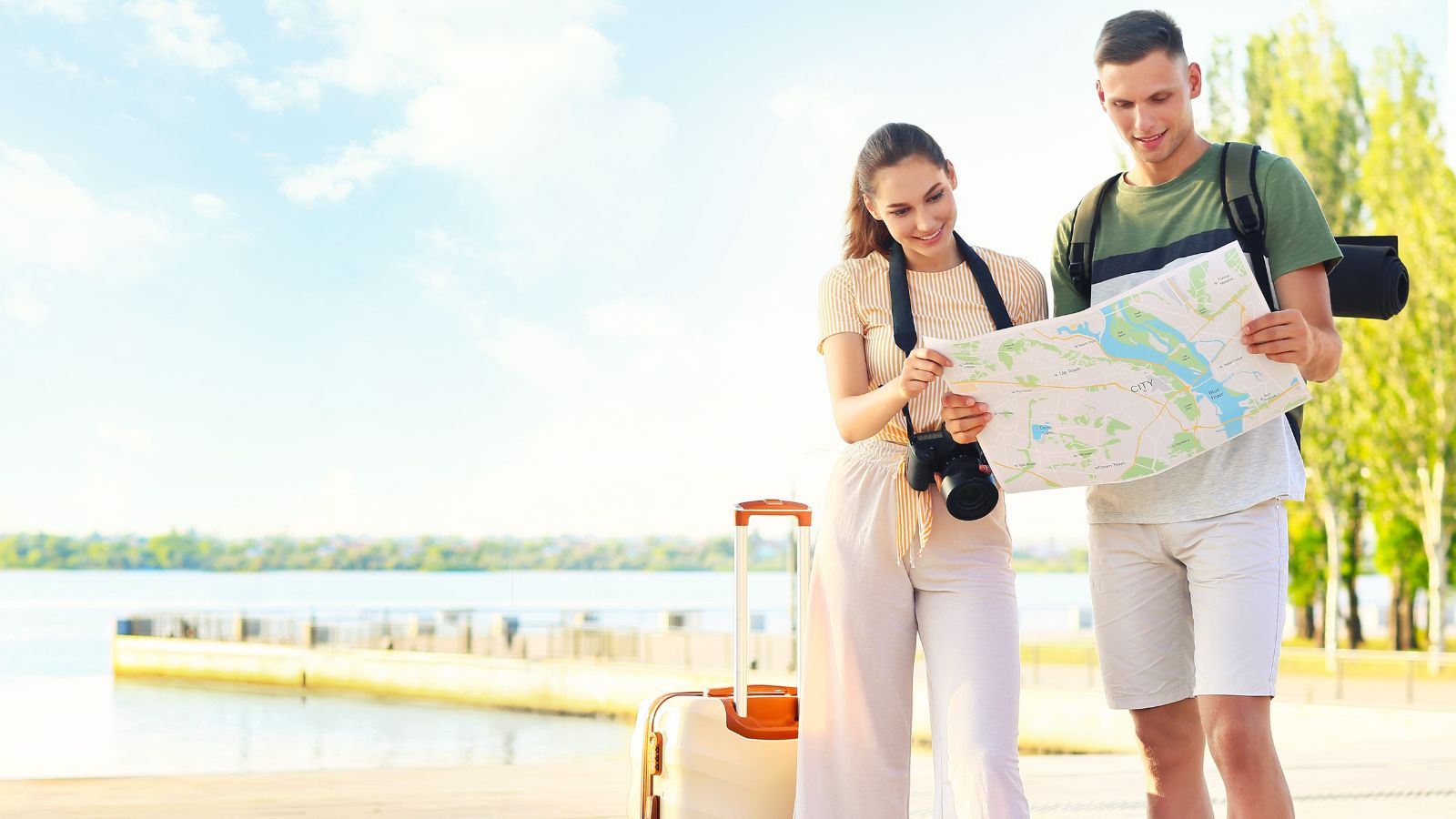 A man and woman by the water look at a map, with a camera on her neck, suitcases beside them, and trees under a bright sky.