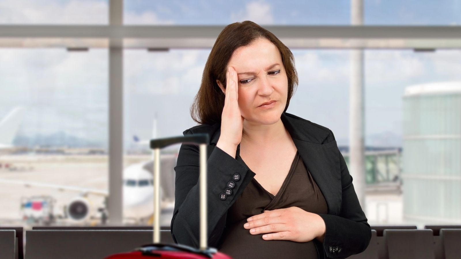 A woman in an airport terminal with a suitcase holds her head and stomach, looking unwell or stressed.