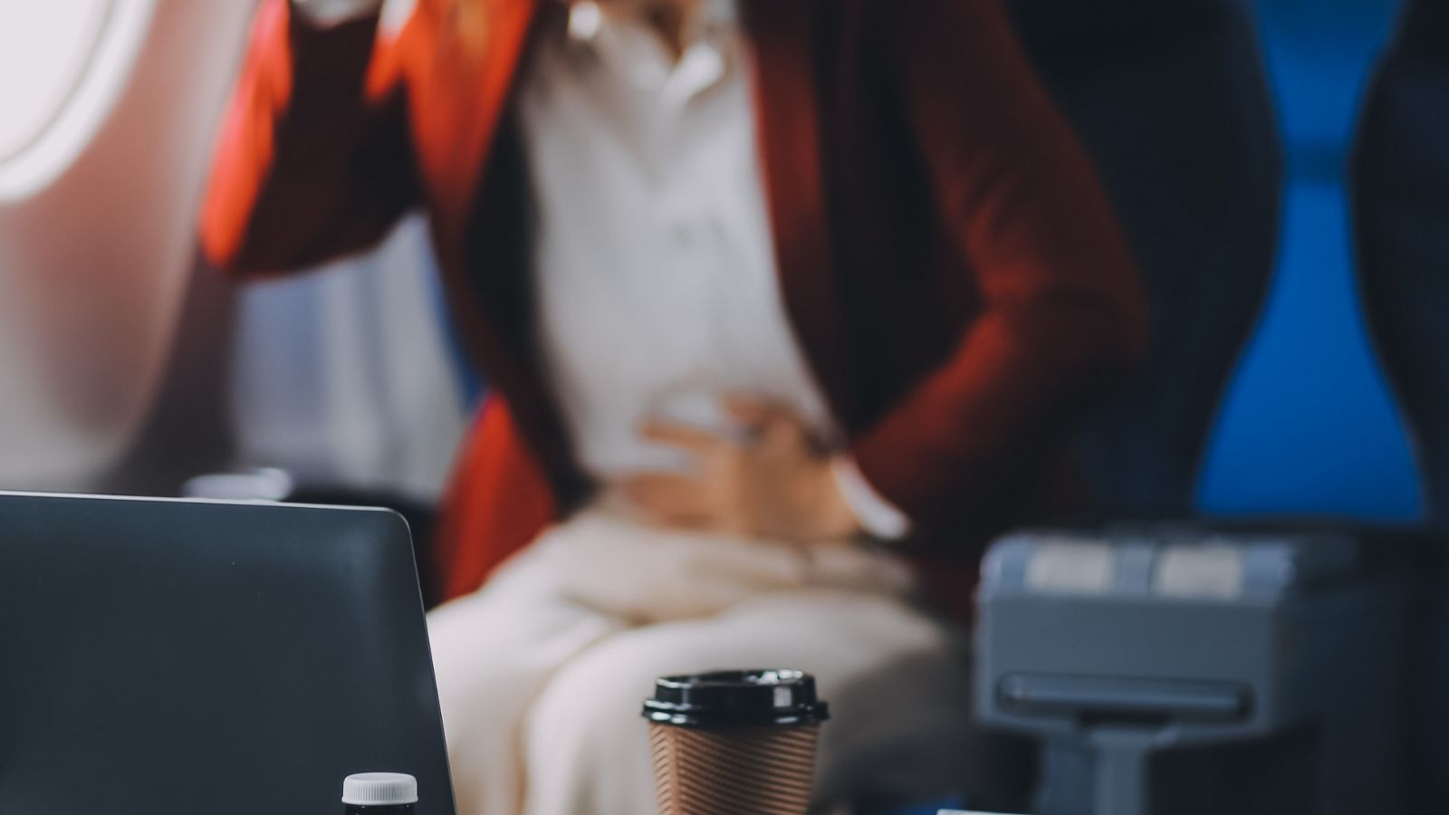 A person in a red jacket on an airplane holds their stomach, with a laptop and coffee cup on the tray table ahead.