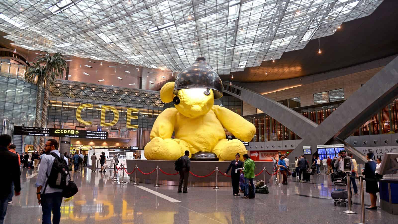 Large yellow teddy bear sculpture with a black lamp on its head stands in an airport terminal atrium as travelers walk past.