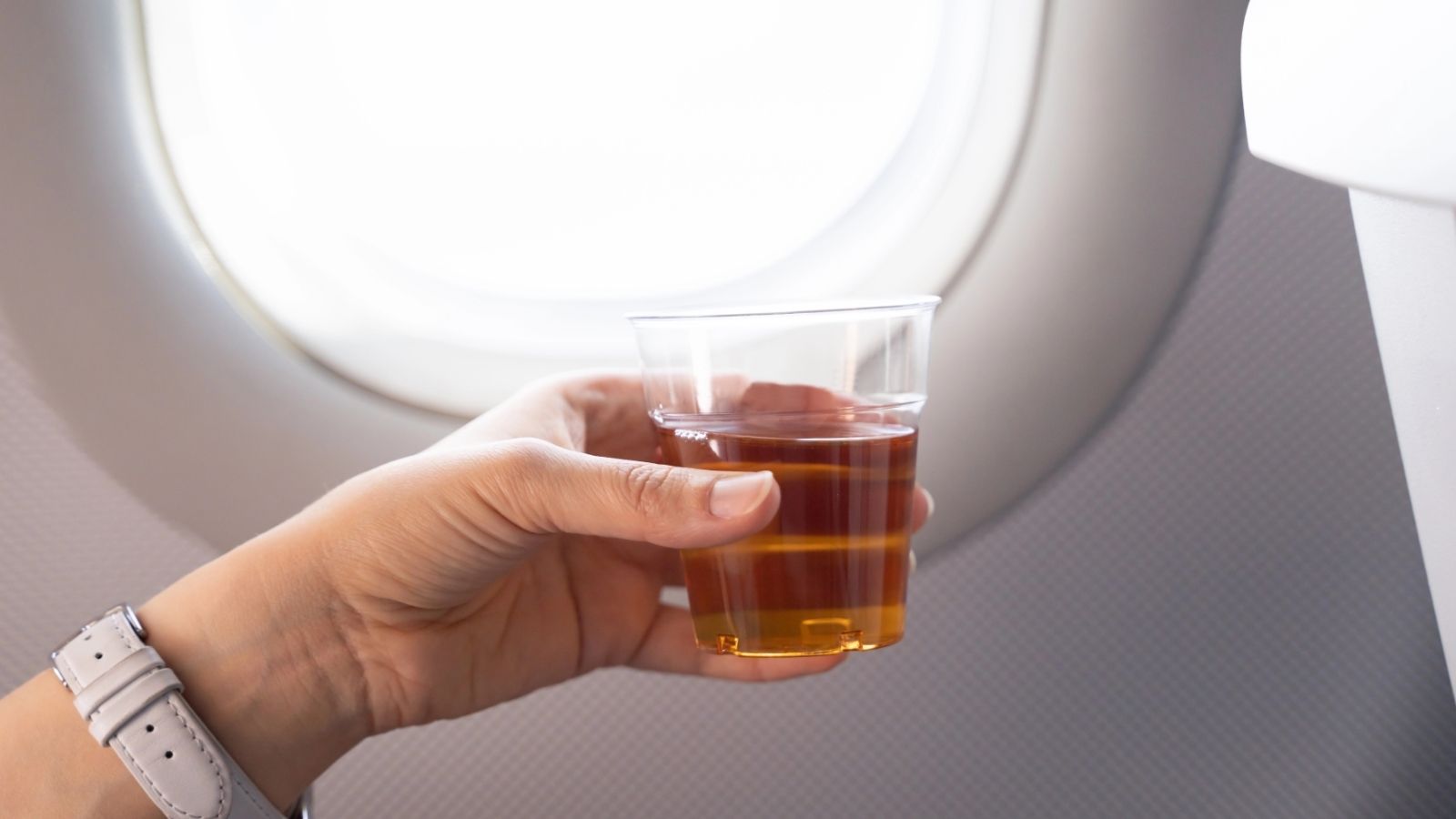 A person holds a clear cup of brown drink by an airplane window.