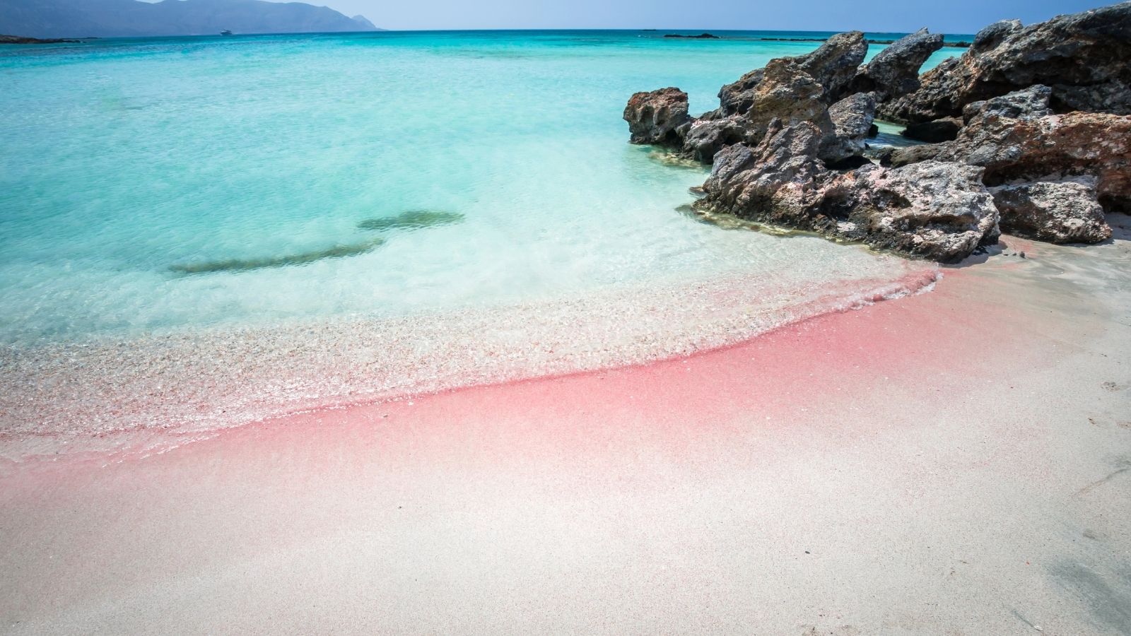 Pink sand shoreline with clear shallow water and rocks at Elafonissi Beach in Crete.