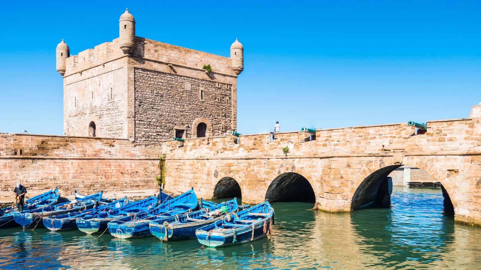 A stone fortress with round turrets stands beside a stone bridge over water. Several blue wooden boats are moored in the foreground under a clear blue sky.