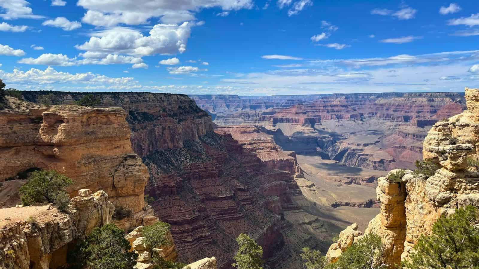 A panoramic view of the Grand Canyon under a bright blue sky with scattered clouds. The scene features steep, rocky cliffs with layers of red, orange, and brown, alongside patches of green vegetation.