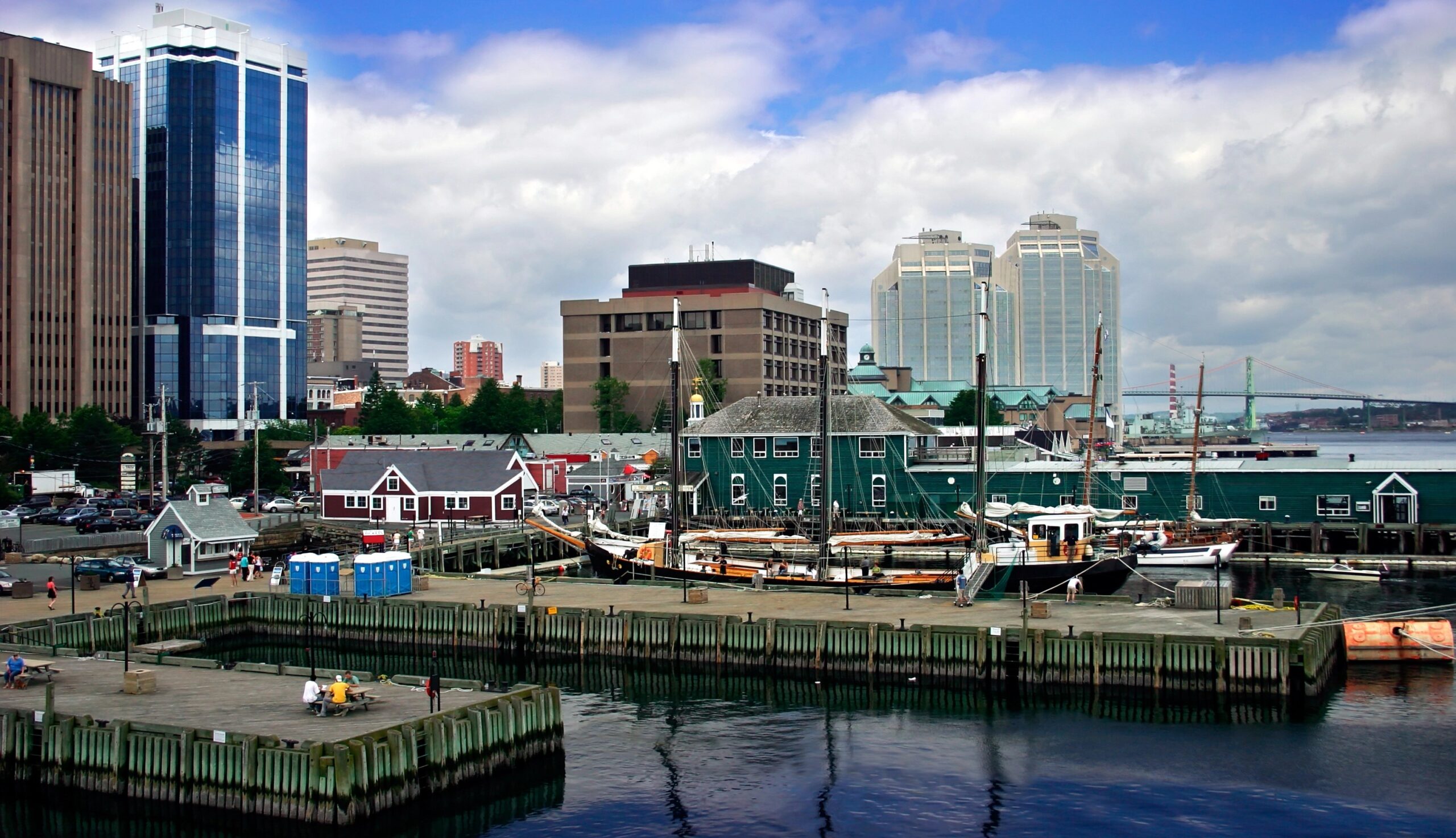 Boats dock at a harbor beside green buildings, with tall city structures and a partly cloudy sky in the background.