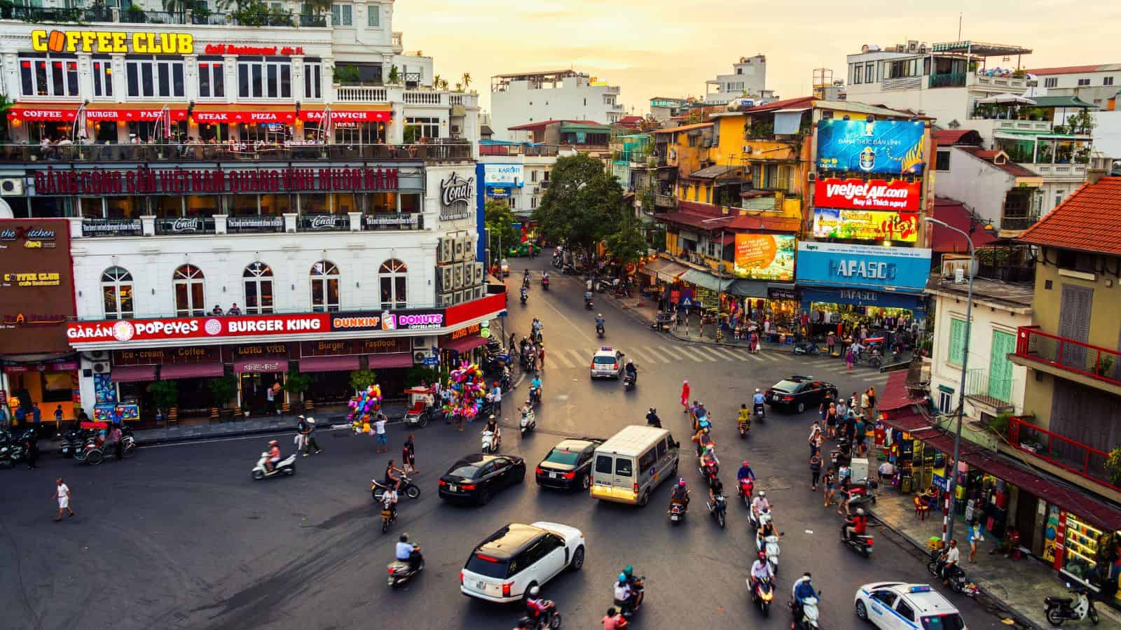 Crowded city intersection at sunset with vehicles, people, and multi-story buildings housing Burger King, Popeyes, and cafes.