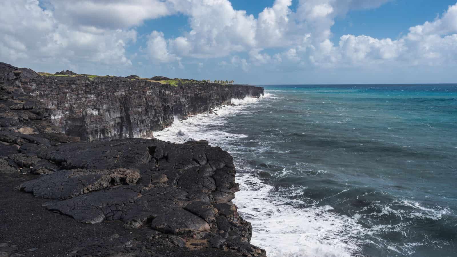 A rocky coastline stretches under a partly cloudy sky. Dark volcanic rocks line the shore, with waves crashing against them. The ocean transitions from deep blue to turquoise closer to the shore, and green foliage is visible atop the cliffs in the distance.