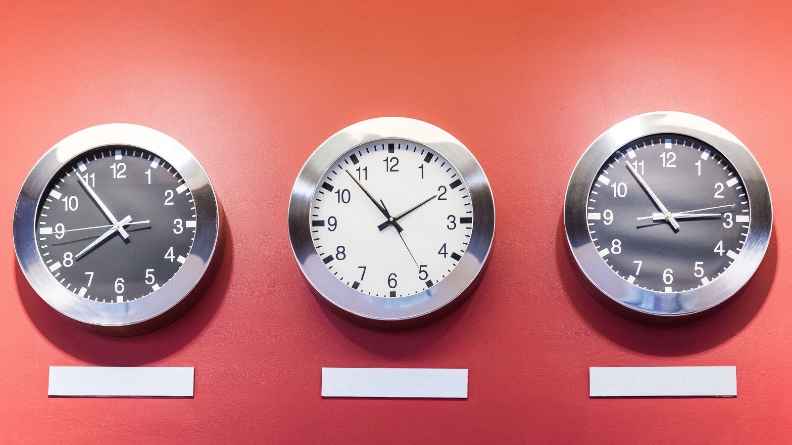 Three silver-framed wall clocks showing different times hang on a red wall above blank labels.