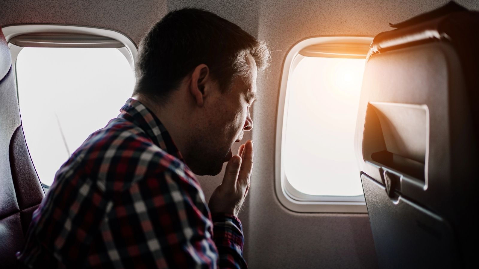 A man in a plaid shirt sits on a plane, hand over mouth, gazing out the window at sunlight.