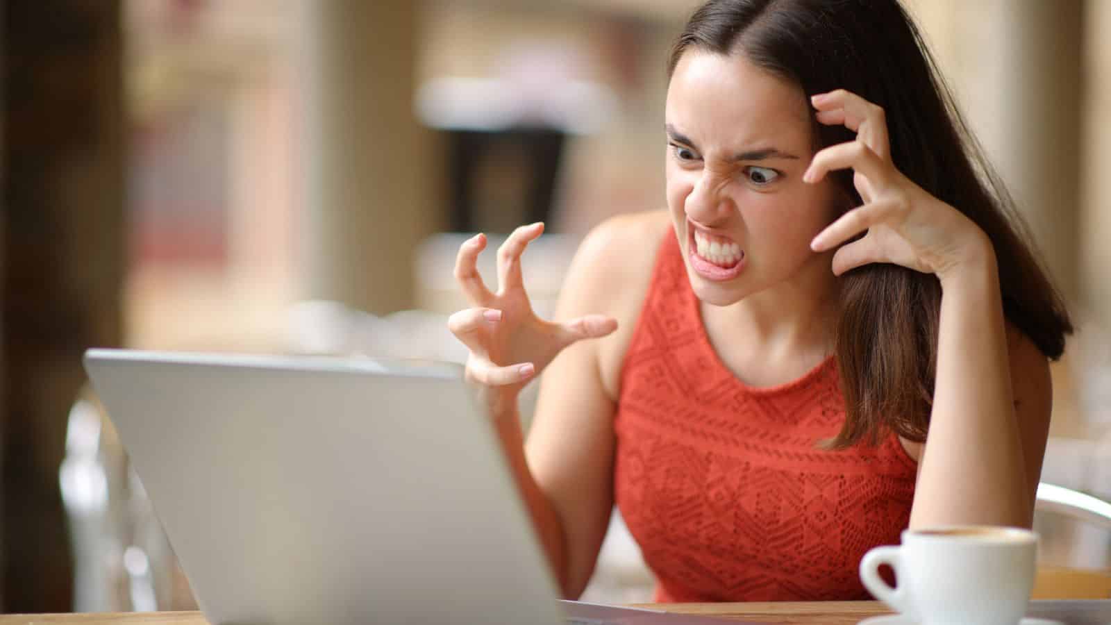 A frustrated woman in an orange top glares at her laptop, clenching her hands in anger beside a coffee cup.