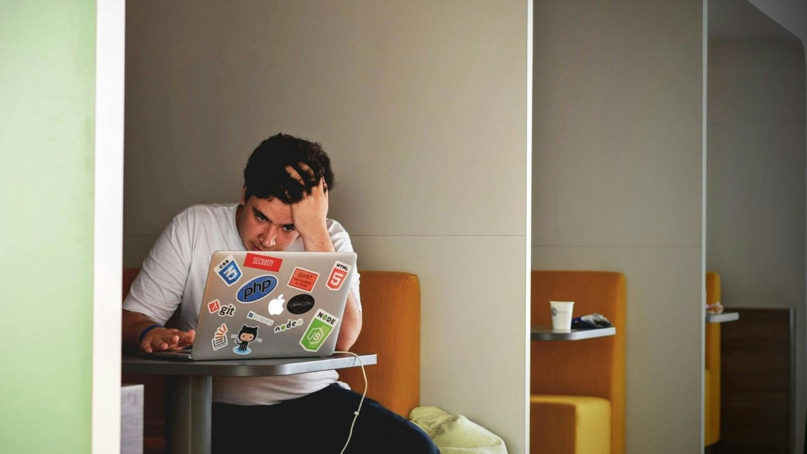 A stressed person works on a programming sticker-covered laptop, holding their head with one hand at a table.
