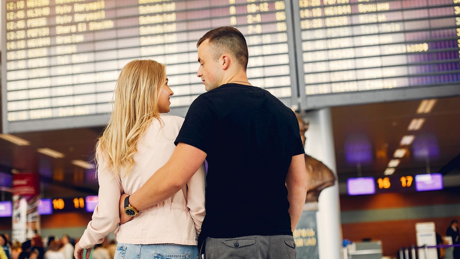 A man and woman in an airport terminal look at a large departures board showing flight information.