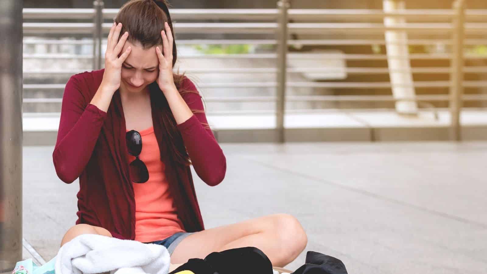 A woman sits outdoors on the ground, hands on her head, gazing down at a pile of clothes in front of her.