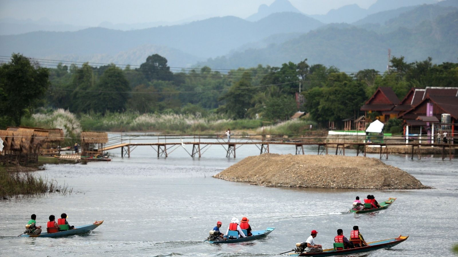 People in life jackets paddle long boats on a river with a wooden footbridge, houses, trees, and mountains in the background.