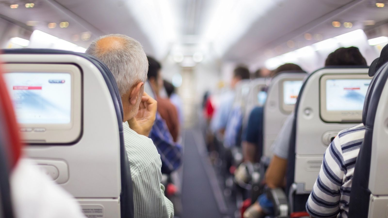 Passengers seen from behind inside an airplane, with seat-back screens and a central aisle visible.