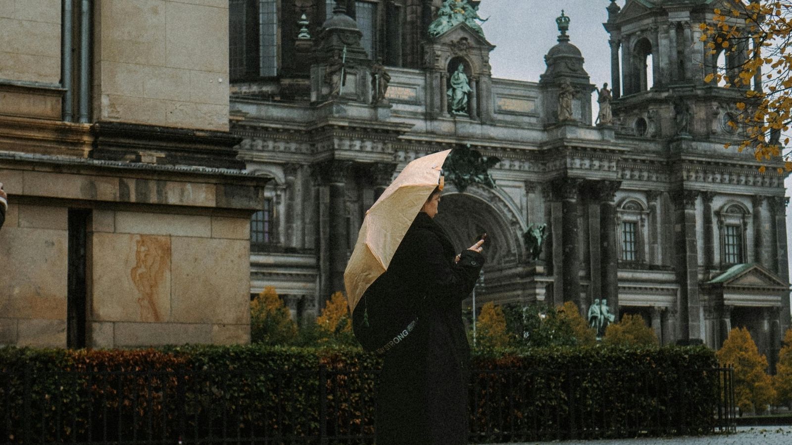 A person with an umbrella stands before an ornate historic building, checking their phone on a cloudy day.