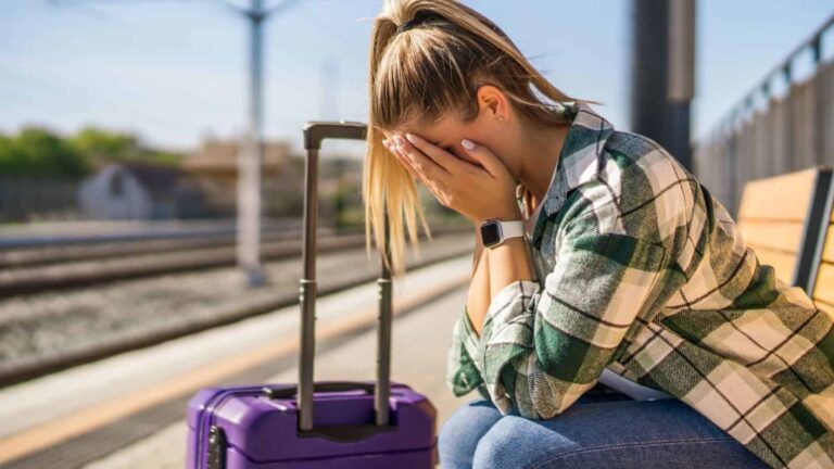 A woman with her head in her hands sits on a bench at a train station beside a purple suitcase.