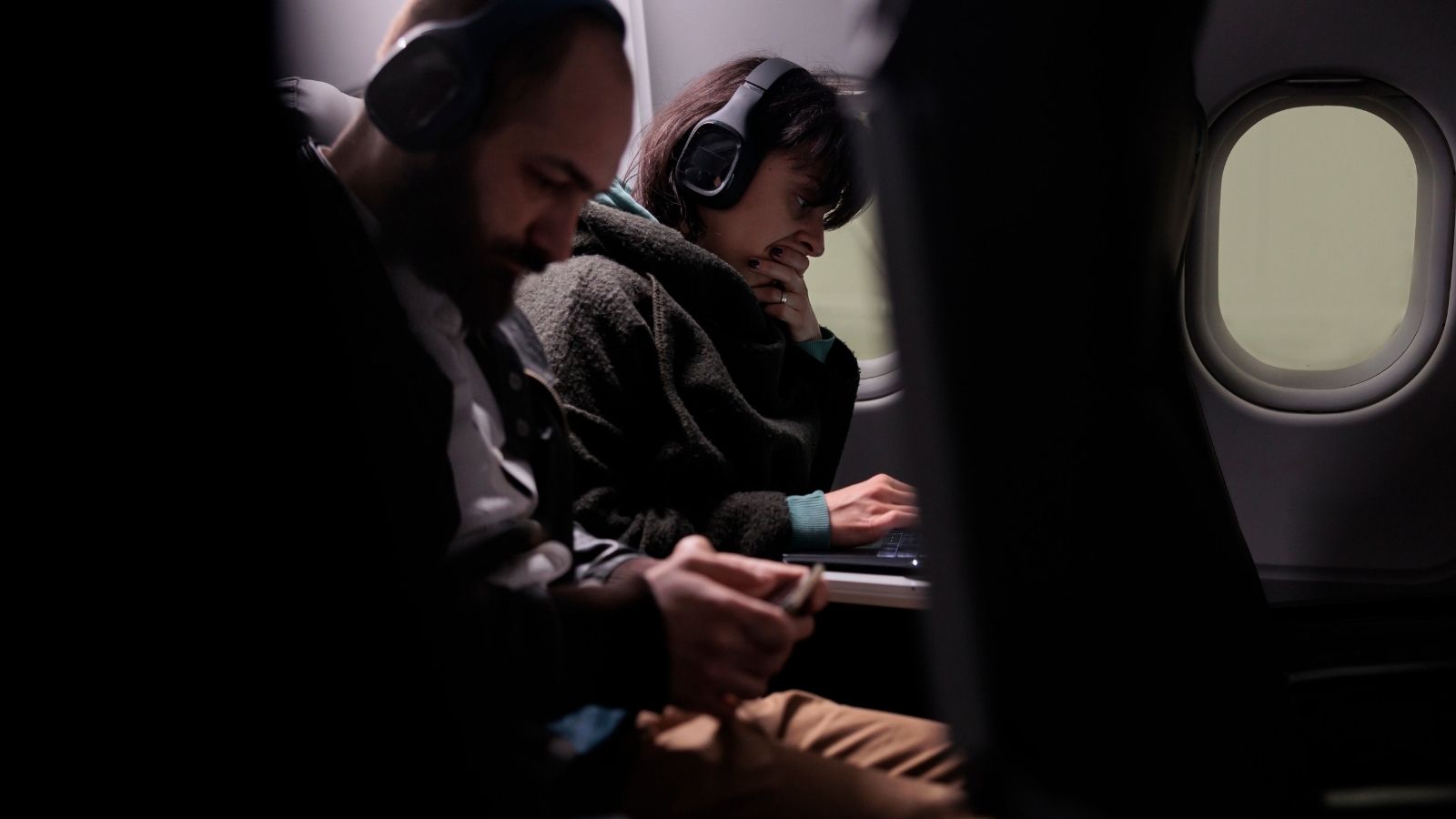Two headphone-wearing airplane passengers sit together, one using a handheld device and the other typing on a laptop by the window.