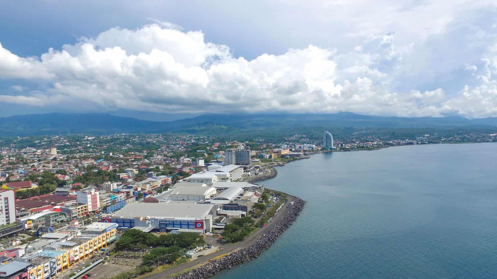 A coastal city with numerous buildings along the shoreline, a curved breakwater, and green hills in the background under a cloudy sky. The water is calm and there are some high-rise structures near the coast.