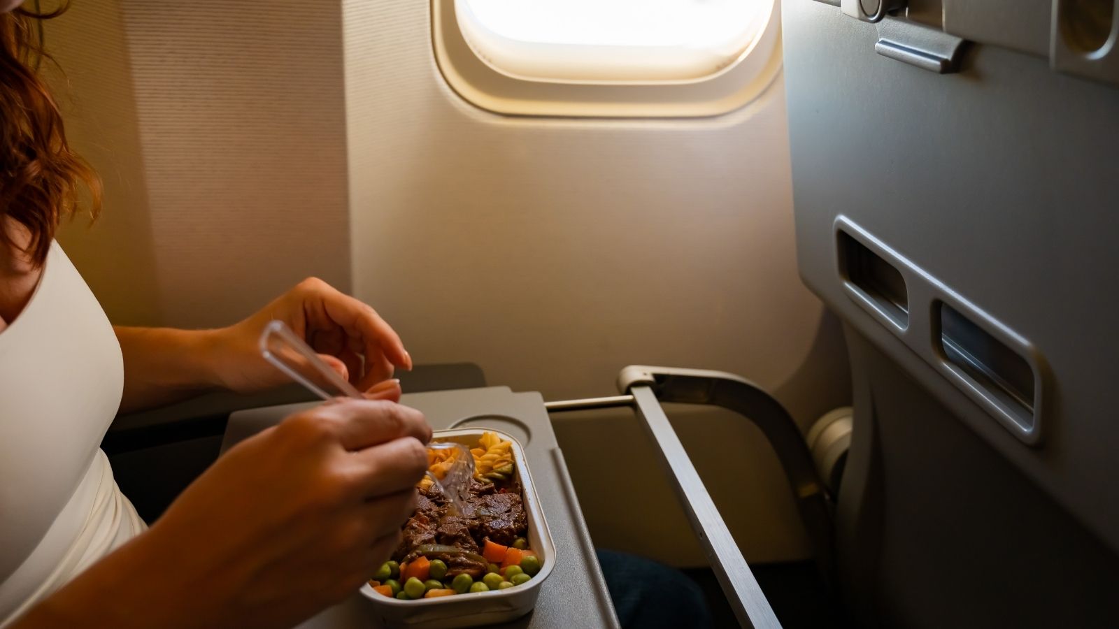 A person eats an airline meal on a tray table by the window during a flight.