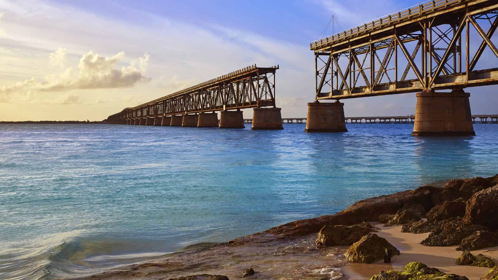 A metal railway bridge extends over clear blue water, partially broken with sections missing. The sky is partly cloudy, and sunlight illuminates the water and rocky shoreline in the foreground.