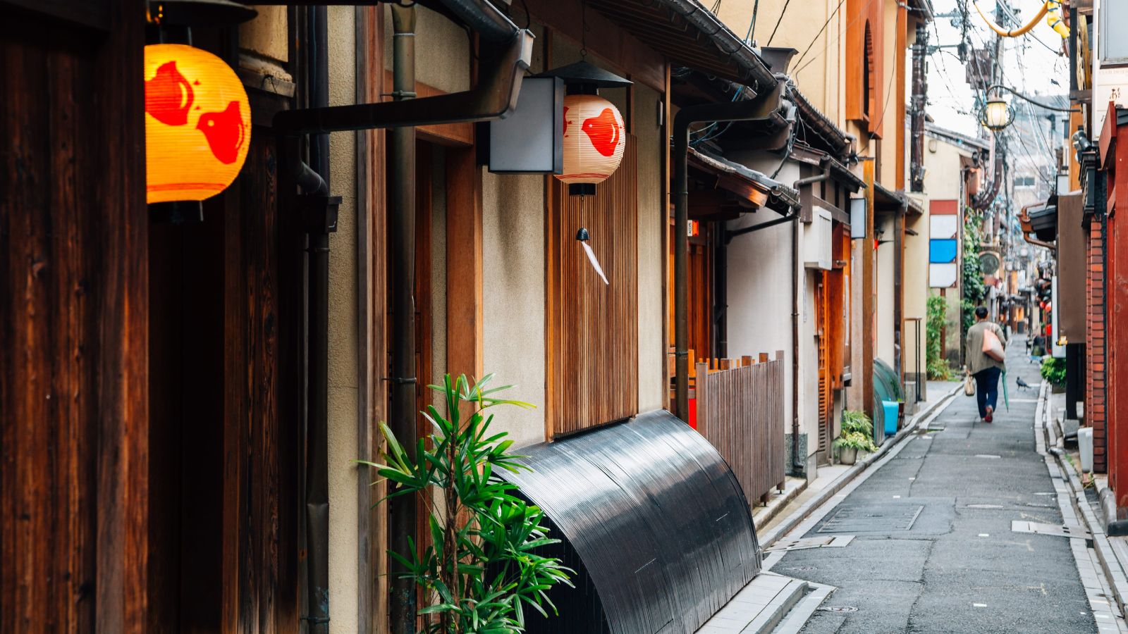 A person walks away down a narrow street lined with traditional Japanese buildings and paper lanterns.