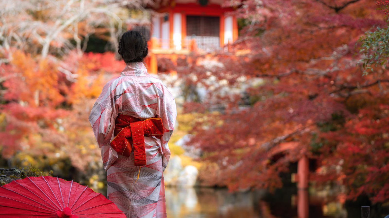 A person in a kimono stands by a red umbrella, facing autumn trees and a traditional Japanese building.