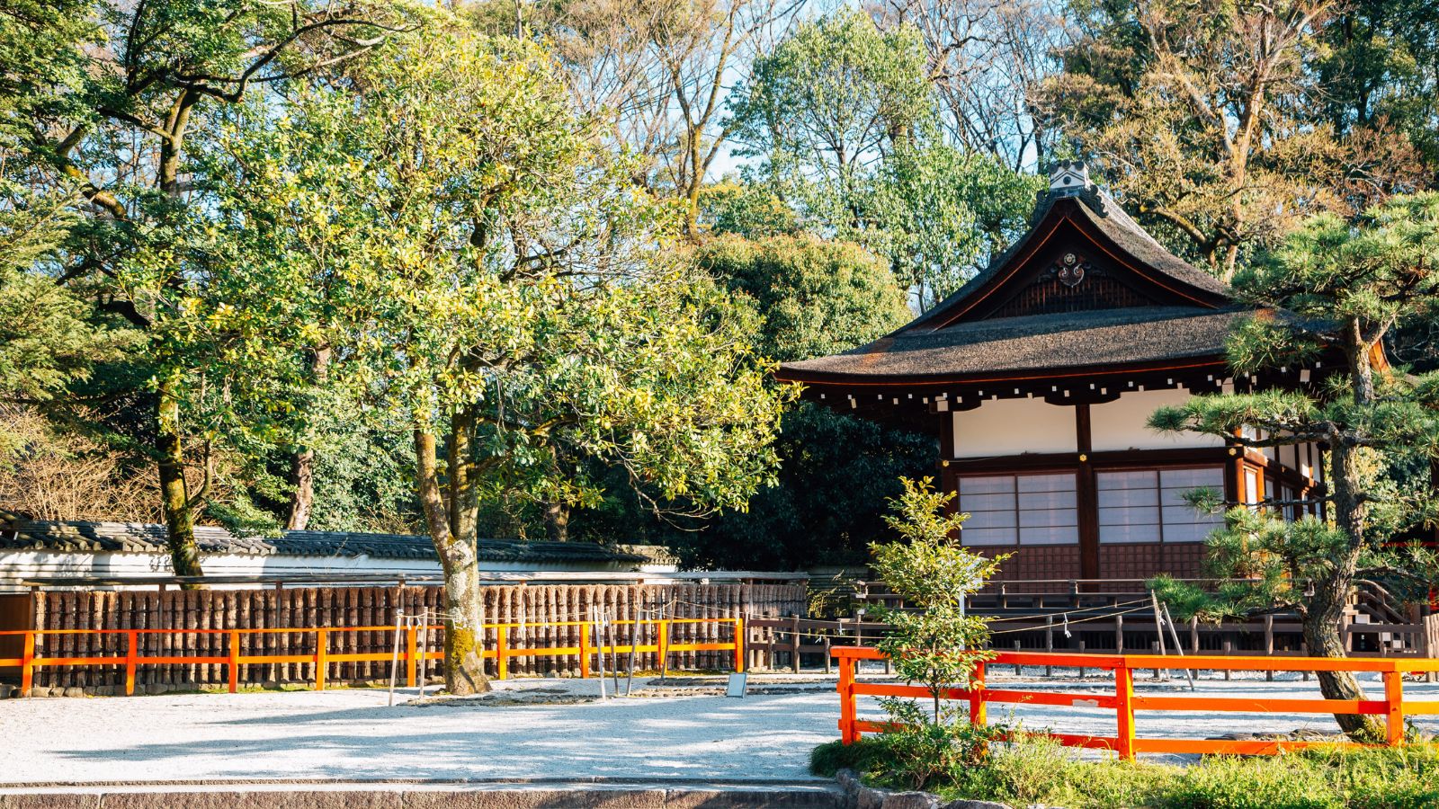 A traditional Japanese wooden building sits by a gravel garden and trees, within red and yellow fences, under a clear sky.