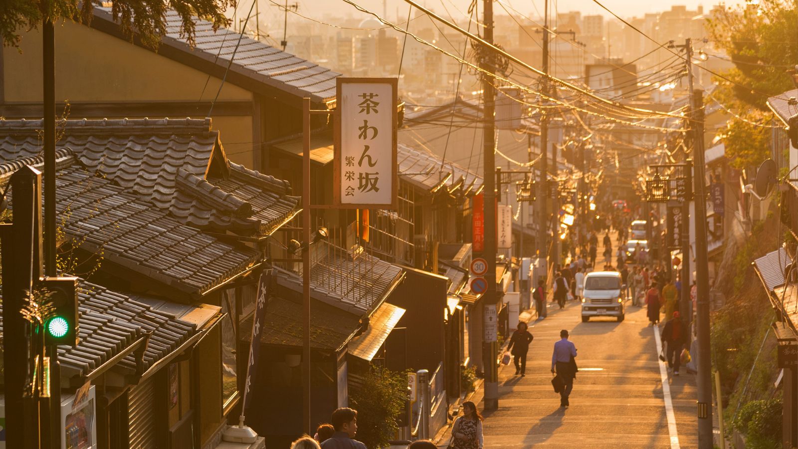 Late-afternoon sunlight illuminates a Japanese street with traditional buildings, hanging signs, pedestrians, and some cars.