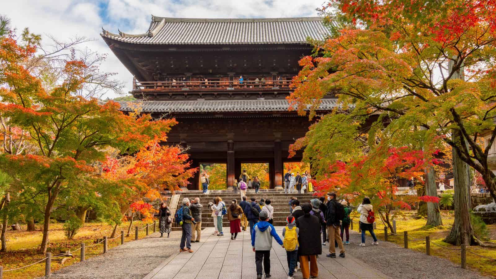 People visit a traditional Japanese temple surrounded by colorful autumn trees and foliage.