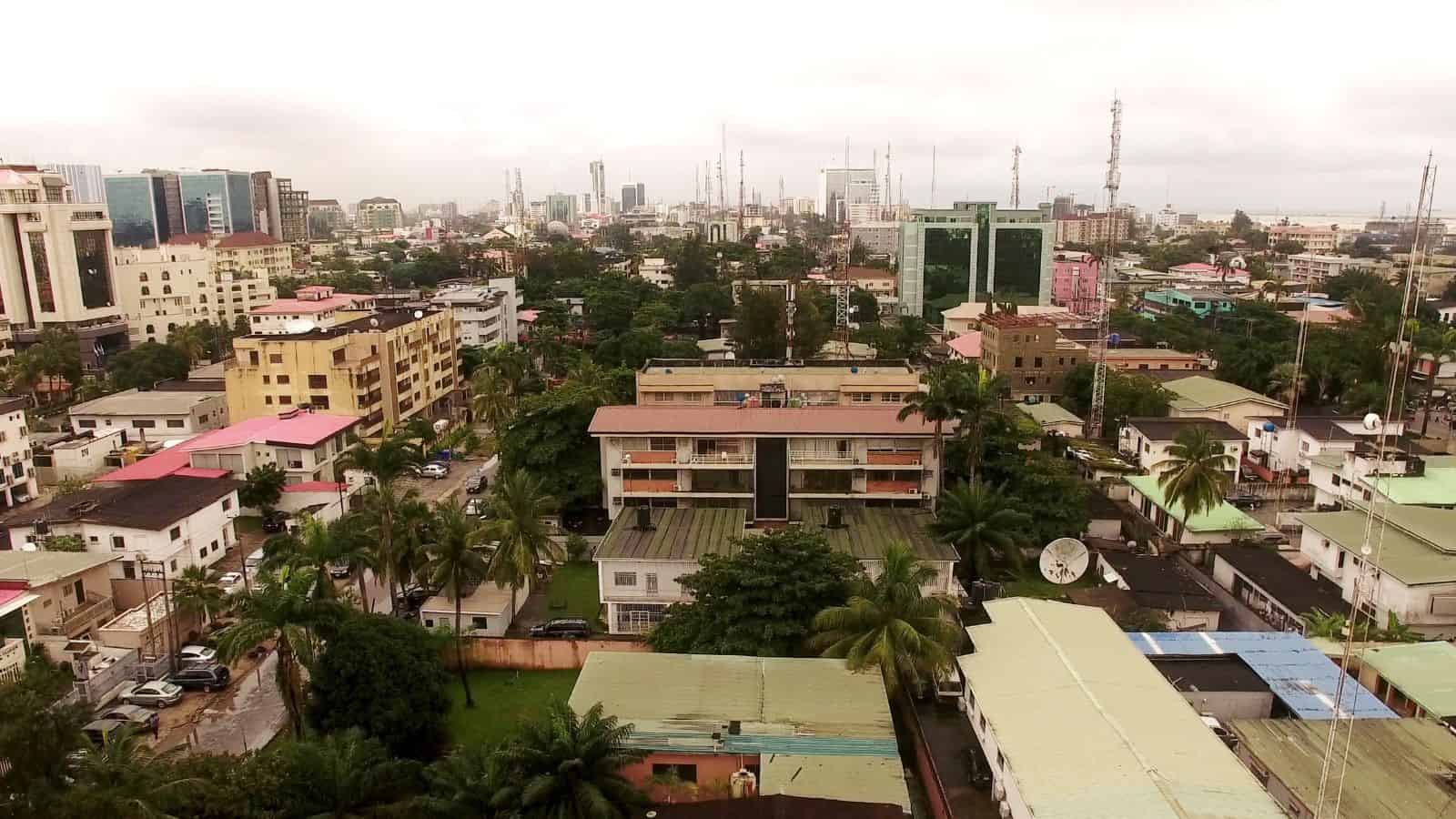 Aerial view of a cityscape featuring a mix of residential and commercial buildings with varied rooftops. The scene includes numerous palm trees, streets with parked cars, and taller buildings in the distant background under a cloudy sky.