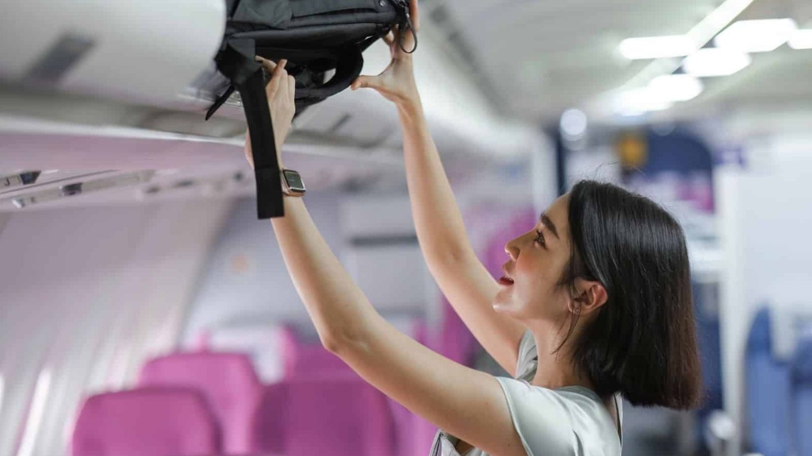 A woman puts a black bag in an overhead compartment inside an airplane cabin with pink seats.