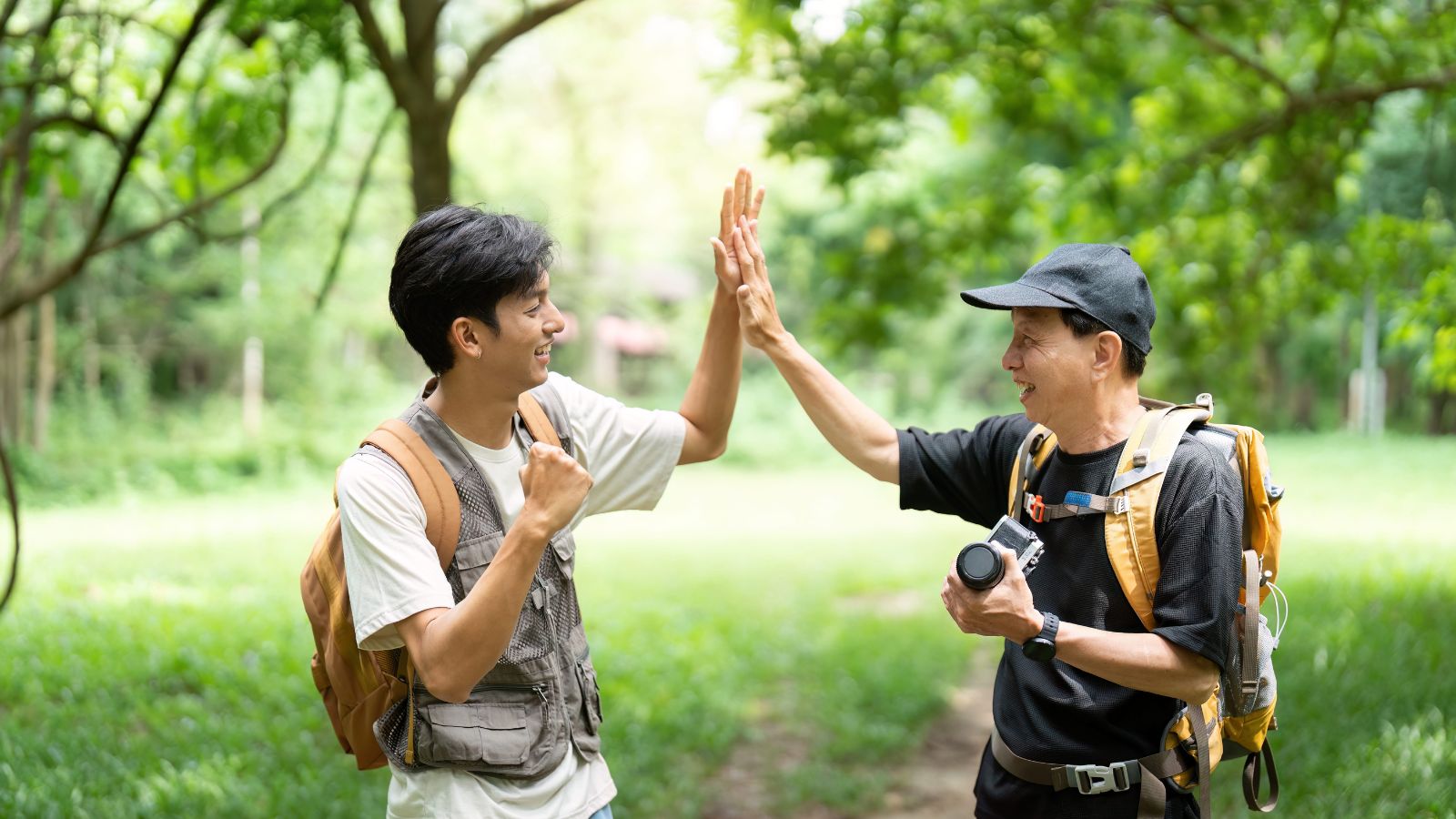 Two people in outdoor gear and backpacks high-five on a grassy path among trees, one holding a camera.