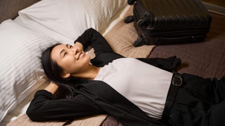 Smiling woman in business attire relaxing on a hotel bed with a suitcase beside her.
